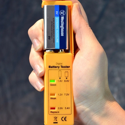 Hand holding yellow digital battery tester with two batteries inserted and voltage scale