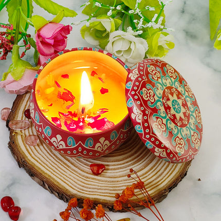 Lit votive tin soy candle with orange wax and scattered petals; patterned lid open
